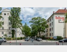 Straßenszene in einem ruhigen Wohngebiet mit mehrstöckigen Gebäuden im Hintergrund, links und rechts flankiert von Bäumen. Auf der rechten Gebäudefassade ist ein großes Logo der „Berliner Baugenossenschaft“ zu sehen, während Autos am Straßenrand geparkt sind und ein Radfahrer die Kreuzung überquert.