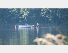 Eine Szene an einem See mit einer Gruppe von Personen, die neben einem kleinen Motorboot im flachen Wasser stehen oder knien. Im Hintergrund befindet sich ein dichter Wald, der bis an das Ufer reicht.
