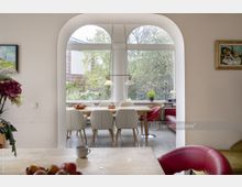 Heller Essbereich mit gebogenem Durchgang und großen Fenstern, die auf eine grüne Außenlandschaft blicken. Ein Holztisch mit weißen Stühlen in der Mitte, umgeben von roten Sesseln und Dekorationen wie Blumen und Obstschalen.