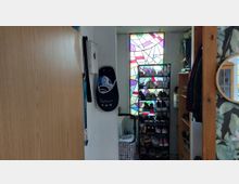 Schmaler Eingangs- bzw. Flurbereich mit einem hohen Schuhregal und Garderobenablage vor einem großen, bunt mosaikartigen Fenster. Links eine helle Holztür und ein Wandhaken, rechts ein offenes Regal und ein runder Spiegel an einer gemusterten Tapete.