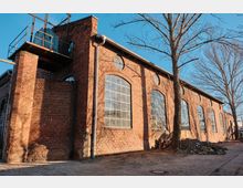 Exterior of a long red-brick industrial building with tall arched multi-pane windows and small circular windows beneath the roofline. A metal platform and water tank sit on a brick tower at the corner, with leafless trees and a paved walkway alongside the building.