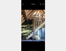 Ein Innenraum mit einer offenen Holzbalkendecke, einer Holztreppe mit rustikalen Geländern und einem Wandbild, das eine Landschaft zeigt. Im Hintergrund befindet sich eine moderne Küche mit weißen und metallischen Elementen.