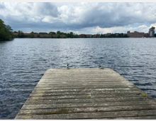 Holzsteg am Ufer eines großen Sees, der in das Wasser hinausragt. Am gegenüberliegenden Ufer sind Bäume und eine städtische Bebauung mit mehrstöckigen Gebäuden unter einem wolkigen Himmel zu sehen.