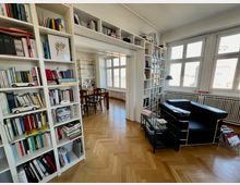 Helles Wohnzimmer mit Bücherregalen, die eine Wand ausfüllen, einem schwarzen Ledersessel und einem Glastisch. Der Raum hat große Fenster, Parkettboden und im Hintergrund ist ein Esstisch mit Stühlen in einem angrenzenden Bereich sichtbar.