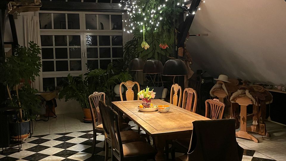 Großer Essbereich in einem loftartigen Raum mit schwarz-weiß kariertem Fliesenboden, langem Holztisch und gemischten Stühlen. Hohe Sprossenfenster, sichtbare Balken und eine Reihe von Hüten unter der Decke, dazu Pflanzen und Lichterketten als Dekoration.