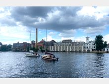 Industriearchitektur am Ufer eines Flusses mit einer alten Brauerei, erkennbar an der hohen Schornsteinsilhouette und Gebäuden mit großen Rundbogenfenstern; im Vordergrund liegen zwei Segelboote auf dem Wasser.