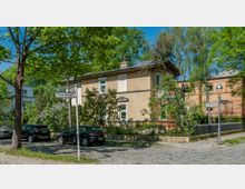 Ecke einer ruhigen Wohnstraße mit Kopfsteinpflaster, umliegenden Bäumen und einem zweistöckigen Haus aus beigem Backstein mit weißen Fenstern. Im Vordergrund sind Straßenschilder und parkende Autos zu sehen.