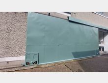 Grüne Metallwand eines Industriegebäudes mit grob verputzter Fassade und einer Schiene am Boden. Rechts sind Teile eines Gebäudeeingangs mit Fenstern sichtbar.