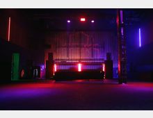 Dunkler Raum eines Clubs oder Veranstaltungsortes mit futuristischer Beleuchtung in Rot- und Violetttönen. Im Hintergrund befindet sich eine Bühne mit Holzpaneelen und seitlich aufgestellten Lautsprechern, die von LED-Lichtern umrahmt sind.