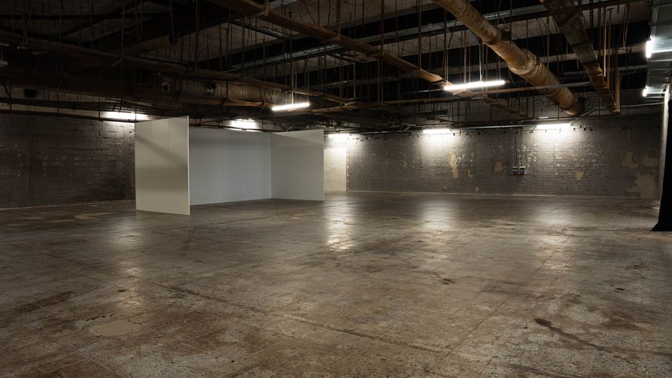 Große, leere Industriehalle mit Betonboden und dunklen Ziegelwänden, in der Mitte stehen mehrere freistehende weiße Stellwände. An der offenen Decke sind sichtbare Rohrleitungen, Stahlträger und Deckenleuchten angebracht.