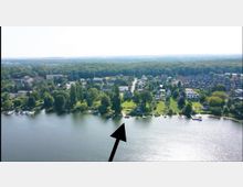 Luftaufnahme einer Stadtlandschaft mit einer Uferpromenade an einem See. Am Ufer befinden sich grüne Flächen, vereinzelte Bäume, kleine Stege und Wohnhäuser, während im Hintergrund Häuser und Wald zu sehen sind.
