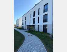 Moderner, weißer Mehrfamilienhauskomplex mit mehreren Stockwerken, großen rechteckigen Fenstern und teils schwarzen Balkongeländern. Ein geschwungener gepflasterter Gehweg führt durch begrünte Vorgärten zum überdachten Hauseingang.