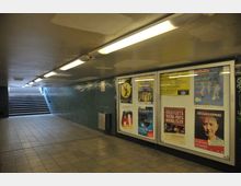 Ein unterirdischer Fußgängertunnel mit gefliesten Wänden in Dunkelgrün und einem beleuchteten Treppenaufgang im Hintergrund. An der rechten Wand hängen mehrere Plakate in Glaskästen, die für Veranstaltungen und Konzerte werben.