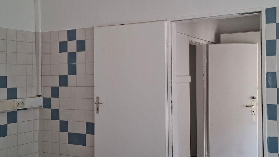 Innenraum mit weiß gekachelten Wänden und blauen Akzentfliesen im Schachbrettmuster sowie einer Steckdosenleiste an der Wand. Rechts sind zwei weiße Türen, eine davon geöffnet, die in einen angrenzenden Raum oder Flur führt.