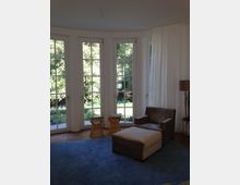 Wohnzimmer mit großen, bodentiefen Sprossenfenstern, die einen Blick in einen Garten bieten. Im Vordergrund stehen ein brauner Sessel mit Fußhocker, zwei kleine Beistelltische und ein blauer Teppich; rechts ist ein Stehlampenfuß erkennbar.