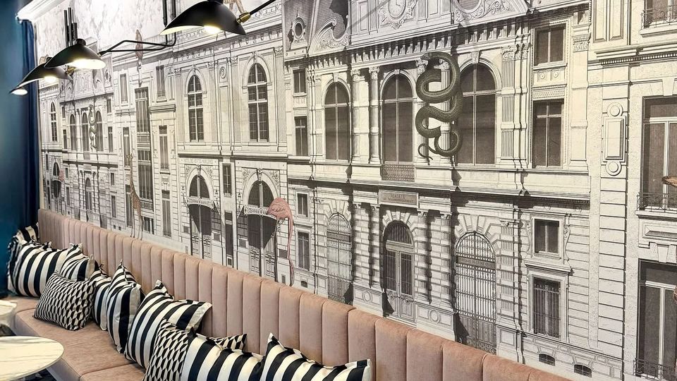 Langer Sitzbereich in einem Café oder Restaurant mit gepolsterten rosa Bänken, gestreiften und geometrisch gemusterten Kissen. An der Wand ist eine großflächige schwarz-weiße Illustration historischer Gebäude zu sehen, beleuchtet von modernen, schwarzen Wandlampen.