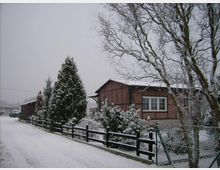 Ein schneebedecktes Dorfhaus aus rotem Backstein mit großen Fenstern und Fachwerkelementen, umgeben von Bäumen, Sträuchern und einem Holzzaun. Schnee liegt auf dem Boden und den Pflanzen, und ein leicht bewölkter Himmel dominiert die Szenerie.