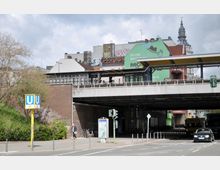 Straßenszene mit einer U-Bahn-Station, deren Eingang unter einer Hochstraße liegt. Im Hintergrund sind Gebäude mit Graffiti und eine an der Fassade montierte Werbetafel sowie ein Kirchturm zu sehen.