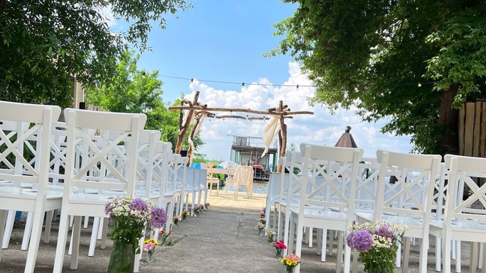 Ein Außenbereich mit einer Zeremonie-Setup: Weiße Stühle sind in Reihen angeordnet, entlang eines Weges mit Blumen in Glasvasen dekoriert. Im Hintergrund ist ein hölzerner Bogen unter Bäumen sichtbar, dahinter ein klarer Himmel und ein Gebäude in der Ferne.