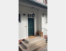 Weiße Hausfassade mit einer dunkelgrünen Tür, die von einem kleinen Vordach mit hängender Laterne überdacht ist. Drei Steinstufen führen zur Tür, daneben steht eine Holzbank, und eine Fußmatte liegt vor dem Eingang.