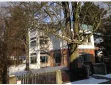 Ein zweistöckiges Wohnhaus mit einer Fassade aus weißem Putz und roten Ziegeln, umgeben von schneebedecktem Boden. Im Vordergrund steht ein großer, unbelaubter Baum, und ein niedriger Zaun grenzt das Grundstück zur Straße ab.