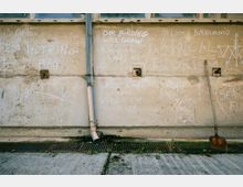 Eine verwitterte Betonwand mit Kreidezeichnungen und -schriftzügen, umgeben von mehreren kleinen quadratischen Wandöffnungen. Ein Fallrohr läuft senkrecht entlang der Wand, und am Boden befinden sich ein Metallgitter mit Abfluss sowie eine Schaufel, die an der Wand lehnt.