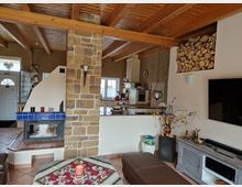 Wohnzimmer mit offener, rustikaler Gestaltung. Zentrales Element ist ein Kamin mit Steinverkleidung und blauer Fliesenumrandung, daneben ein Wandregal mit gestapeltem Feuerholz. Sichtbare Holzbalkendecke, Fliesenboden und Dekoration wie ein Esstisch mit Kerze und Pflanzenschmuck. Im Hintergrund ist eine helle Küche erkennbar.