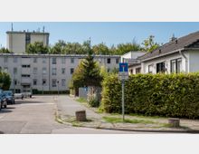 Wohnstraße mit mehrstöckigem Wohngebäude aus hellem Putz links und einem einstöckigen Haus mit dunklem Dach rechts, getrennt durch Hecken. Ein Straßenschild weist auf die Sackgasse „Liviusstraße“ hin, im Hintergrund sind Bäume und ein weiteres hohes Gebäude sichtbar.