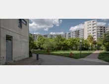Wohngebiet mit mehrstöckigen Plattenbau-Wohnhäusern im Hintergrund, umgeben von Bäumen und Grünflächen. Im Vordergrund führen ein asphaltierter Weg und ein gepflasterter Pfad durch eine parkähnliche Anlage mit Straßenlaternen.