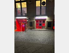 Nachtaufnahme einer Kopfsteinpflasterstraße vor einem Gebäude mit beleuchteten Eingängen. Der Eingang zur "SusPiria Bar" ist mit roten und violetten Akzenten gestaltet, darunter rote Fenster und ein leuchtendes Schild; daneben eine Treppe mit rotem Geländer. Oben befinden sich beleuchtete Fenster und Reklameschilder.