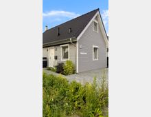 Einfamilienhaus mit grauer Holzfassade und schwarzem Satteldach, umgeben von einem kleinen gepflasterten Hof und niedrigem Bewuchs im Vordergrund. Das Gebäude hat weiße Fensterrahmen und eine Haustür mit integriertem Fenster.