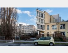 Ein modernes Gebäude mit gelblichen Fassaden, großen Fenstern und abgerundeten architektonischen Details; im Hintergrund ein mehrstöckiger Bürokomplex. Im Vordergrund ein geparkter hellgrüner Kleinwagen, flankiert von Bäumen und niedrigem Gesträuch.