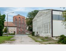 Industriehof mit einem älteren Backsteingebäude im Hintergrund, das mehrere kleine Fenster besitzt, neben einem flachen Wohnhaus mit rotem Ziegeldach. Auf der rechten Seite steht eine große, modernere Lagerhalle mit einer Wand aus Betonplatten. Im Vordergrund ist ein gepflasterter Hof mit etwas Grasbewuchs sichtbar.