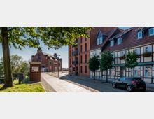 Straßenszene in einer Altstadt mit Kopfsteinpflaster, Fachwerkhäusern mit roten Ziegeldächern und einem Backsteingebäude im Hintergrund. Links im Bild befinden sich Bäume und eine niedrige Mauer mit Blick auf eine grüne Landschaft.