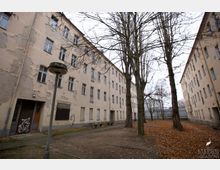 Hinterhof eines alten, mehrstöckigen Wohngebäudes mit bröckelnder Fassade, nackten Bäumen und Laub bedecktem Boden. Im Hof befinden sich eine Laterne, Sitzbänke und ein Gehweg.