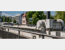 Eine Straßenszene mit industriellen Metallstrukturen im Vordergrund, dahinter mehrere Gebäude, darunter ein modernes Wohnhaus mit Balkonen und ein älteres Haus mit rotem Ziegeldach. Im Hintergrund sind grüne Bäume und ein blauer Himmel sichtbar.