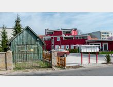 Ein kleiner, grüner Holzschuppen mit verwitterter Oberfläche steht hinter einem Metallzaun. Im Hintergrund befindet sich ein modernes, rot-weißes Wohngebäude mit mehreren Balkonen, ein Hof und ein Briefkastenständer mit der Aufschrift „Florastraße 36“.