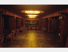 Innenraum eines Veranstaltungsraums mit dunklem Holzboden, roten Wänden und warmer Beleuchtung durch große, ovale Kronleuchter. Im Hintergrund befindet sich eine Bar, umgeben von roten Polsterstühlen und kleinen Tischen, die entlang der Seitenwände verteilt sind.