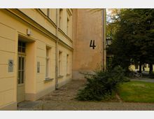 Gelbes, mehrstöckiges Gebäude mit nummeriertem Eingang „3“ und großen Fenstern. Daneben ist eine Gebäudewand mit schwarzer Zahl „4“, während im Hintergrund Bäume, ein Fahrrad und ein gepflasterter Weg zu sehen sind.