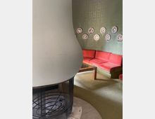 Ein Wohnraum mit grünlich gemusterter Tapete, dekoriert mit einer Reihe von Ziertellern. Im Vordergrund steht ein schlichter, moderner Kamin mit rundem Sockel und Metallgitter. Im Hintergrund befindet sich eine rote Polstereckbank und ein rechteckiger Holztisch auf grünem Teppichboden.