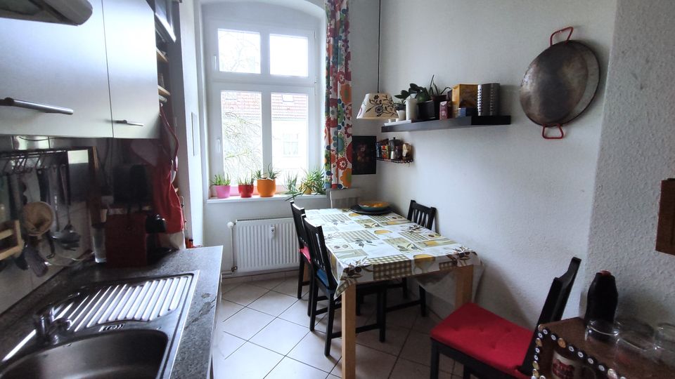 Narrow galley kitchen with a stainless sink and countertop on one side and a small dining table with chairs on the other. A tall arched window with patterned curtains and potted plants brings in light, with wall shelves, hanging cookware, and white tile flooring.