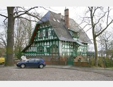 Fachwerkhaus mit grünen und weißen Holzelementen, steilem Schieferdach und rotem Schornstein, umgeben von Bäumen. Im Vordergrund befindet sich eine gepflasterte Straße mit einem geparkten blauen Auto.