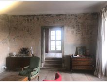 Ein rustikales Wohnzimmer mit abgewitterten Wänden, einem Holzboden und zwei antiken Kommoden. In der Mitte führt eine Türöffnung mit Stufen zu einem Raum mit einem Fenster, durch das Tageslicht einfällt.