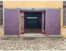 Ziegelwand mit gelber Backsteinfassade, doppeltüriger Eingang mit violetten Holzschiebetüren und Glasflächen, teilweise geöffnet. Vor dem Gebäude ist ein Pflasterboden aus unregelmäßigen Steinen sichtbar.