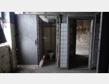 Verlassene, heruntergekommene Toilette in einem dunklen Gebäude mit beschädigten Kacheln, einer offenen Metalltür und einer durch Vandalismus geprägten Umgebung. Im Hintergrund sind Wände mit abblätterndem Putz sichtbar.