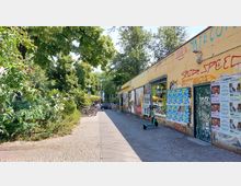 Gepflasterter Fußgängerweg mit links grünen Büschen und Bäumen sowie rechts einer Graffiti-bedeckten gelben Hauswand mit Plakaten. Im Hintergrund sind Fahrräder, eine Sitzmöglichkeit und ein gelbes Sonnendach zu sehen.