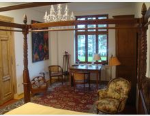 Ein gemütliches Schlafzimmer mit einem Himmelbett aus Holz, einem floralen Sessel, einem orientalischen Teppich und einem Holztisch mit Stuhl vor einer großen Fensterfront. An der Wand hängt ein farbenfrohes abstraktes Gemälde, und ein Kronleuchter hängt von der Decke.