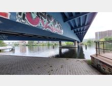 Unterseite einer blauen Brücke mit Graffitis, die über einen Fluss führt. Im Hintergrund sind moderne Gebäude, eine Fußgängerbrücke und grüne Vegetation am Ufer sichtbar.