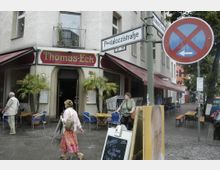 Straßenszene mit dem Eingang eines Restaurants namens „Thomas-Eck“ an einer Ecke, umgeben von Palmen in Töpfen, einer kleinen Außenterrasse mit blauen Stühlen und einem Straßenschild mit der Aufschrift „Pestalozzistraße“. Im Vordergrund gehen Fußgänger vorbei, und auf der rechten Seite ist ein Verkehrsschild mit Park- oder Halteverbot sichtbar.