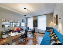 Modern eingerichtetes Büro mit zwei Schreibtischen, Computermonitoren und schwarzen Drehstühlen. Der Raum hat einen bunten Holzboden, helle Wände, große Fenster mit Rollos, mehrere Kunstwerke an den Wänden und ein blaues Sofa mit gemusterten Kissen.